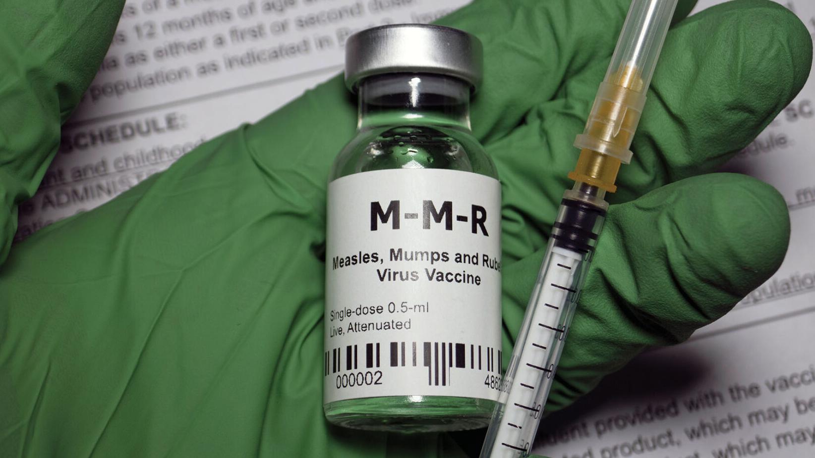 Green medical gloved hand holding a MMR vaccine vial
